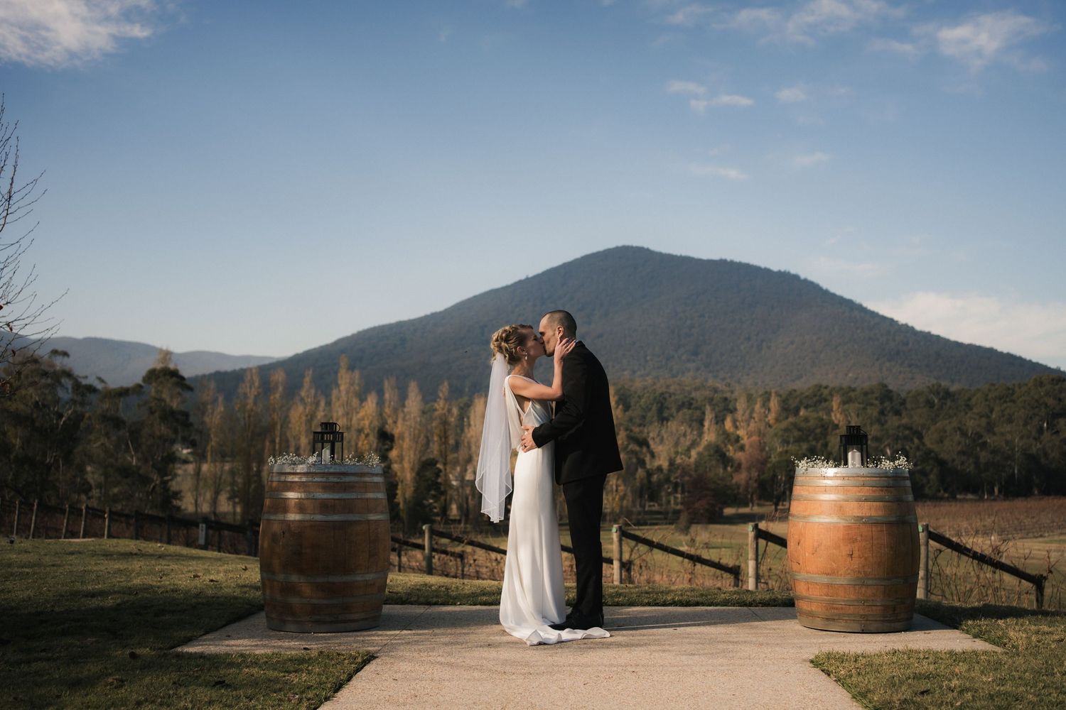 Couple embraces on wedding day at scenic mountain vineyard with rustic wine barrels at sunset at Boat O Craigo, Healesville