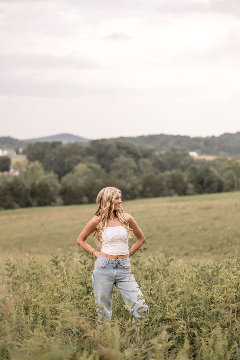 A person in light jeans and crop top poses in a scenic rural meadow with rolling hills in the background.
