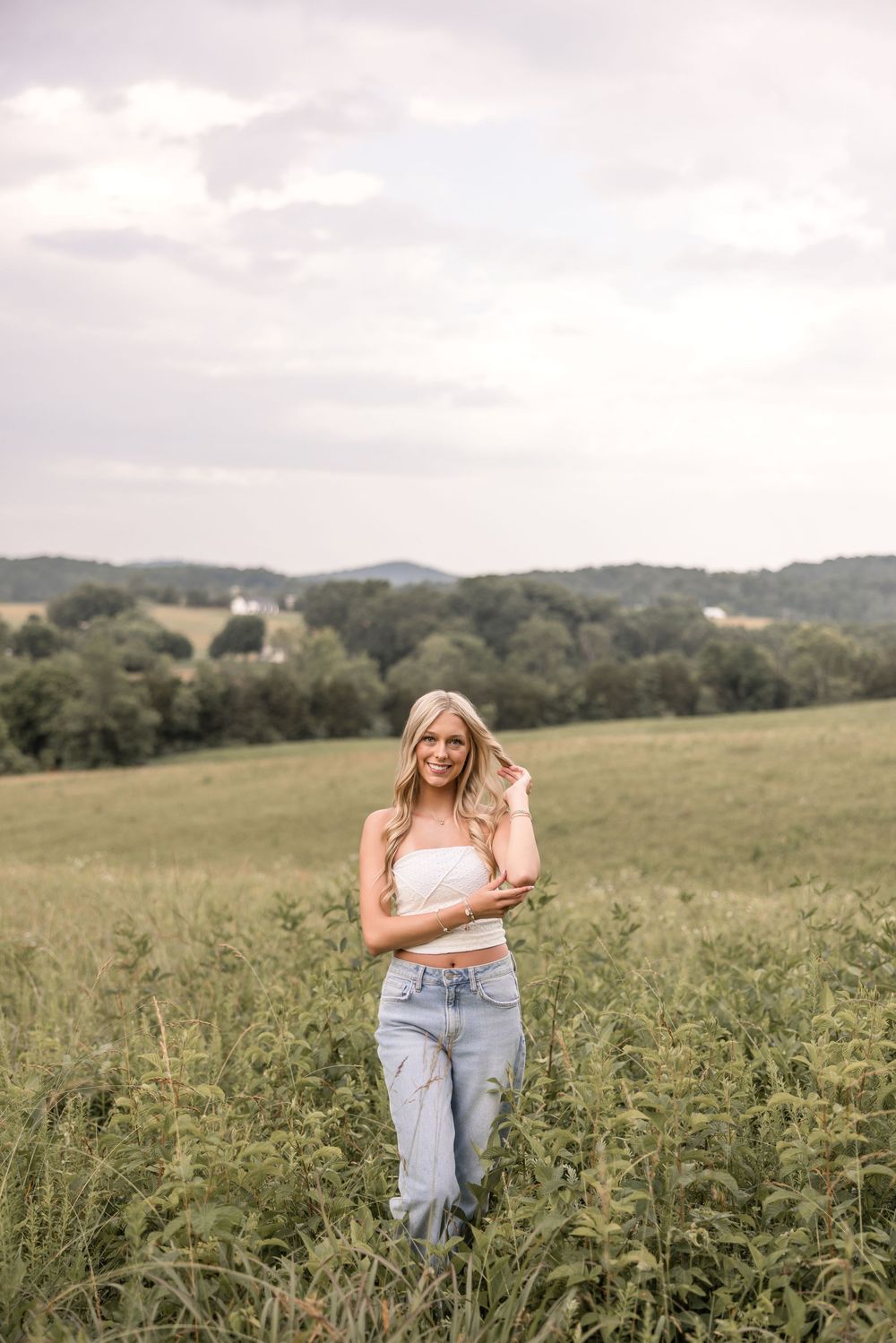 Young woman in casual summer outfit standing in a grassy meadow field with rolling hills in the background at dusk.