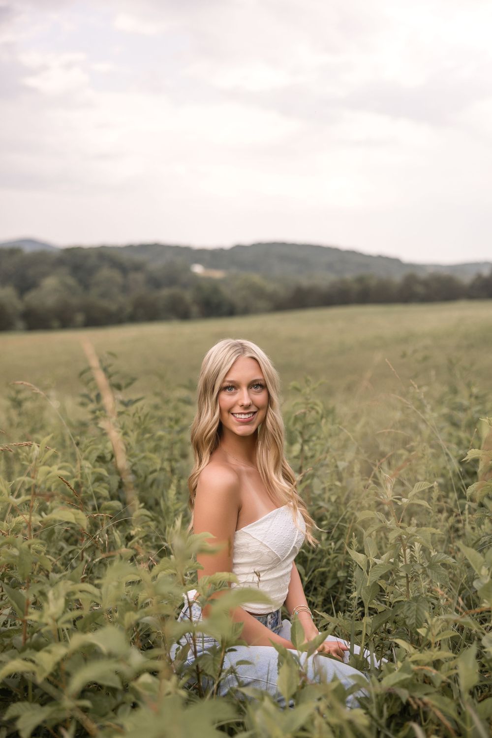 A figure in a white top poses gracefully among tall green plants in a rural field on an overcast summer day.