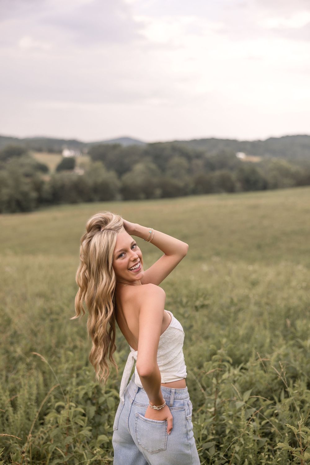 Woman in white top and jeans poses playfully in a scenic meadow with rolling hills in the background.