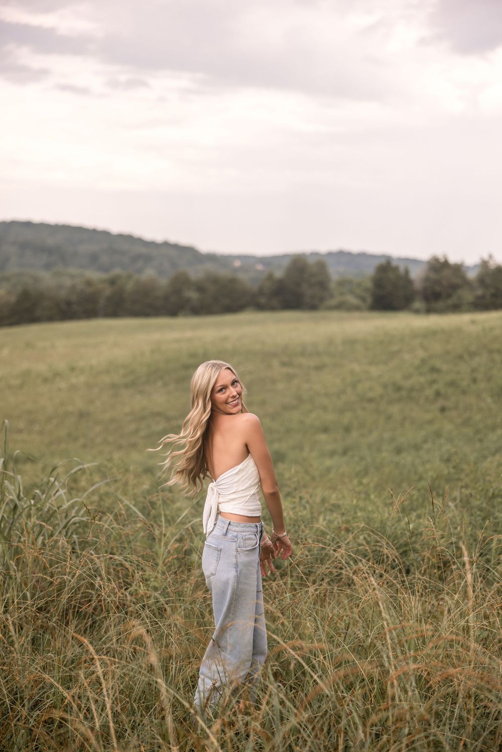 Person in a white top and light jeans stands in tall grass of a pastoral meadow with rolling hills in the background.