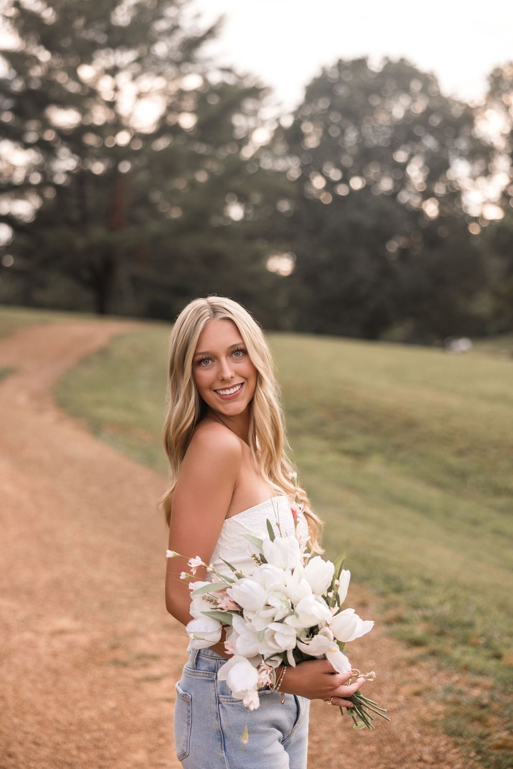 Woman in casual attire holds a beautiful white floral bouquet while standing on a winding dirt path.