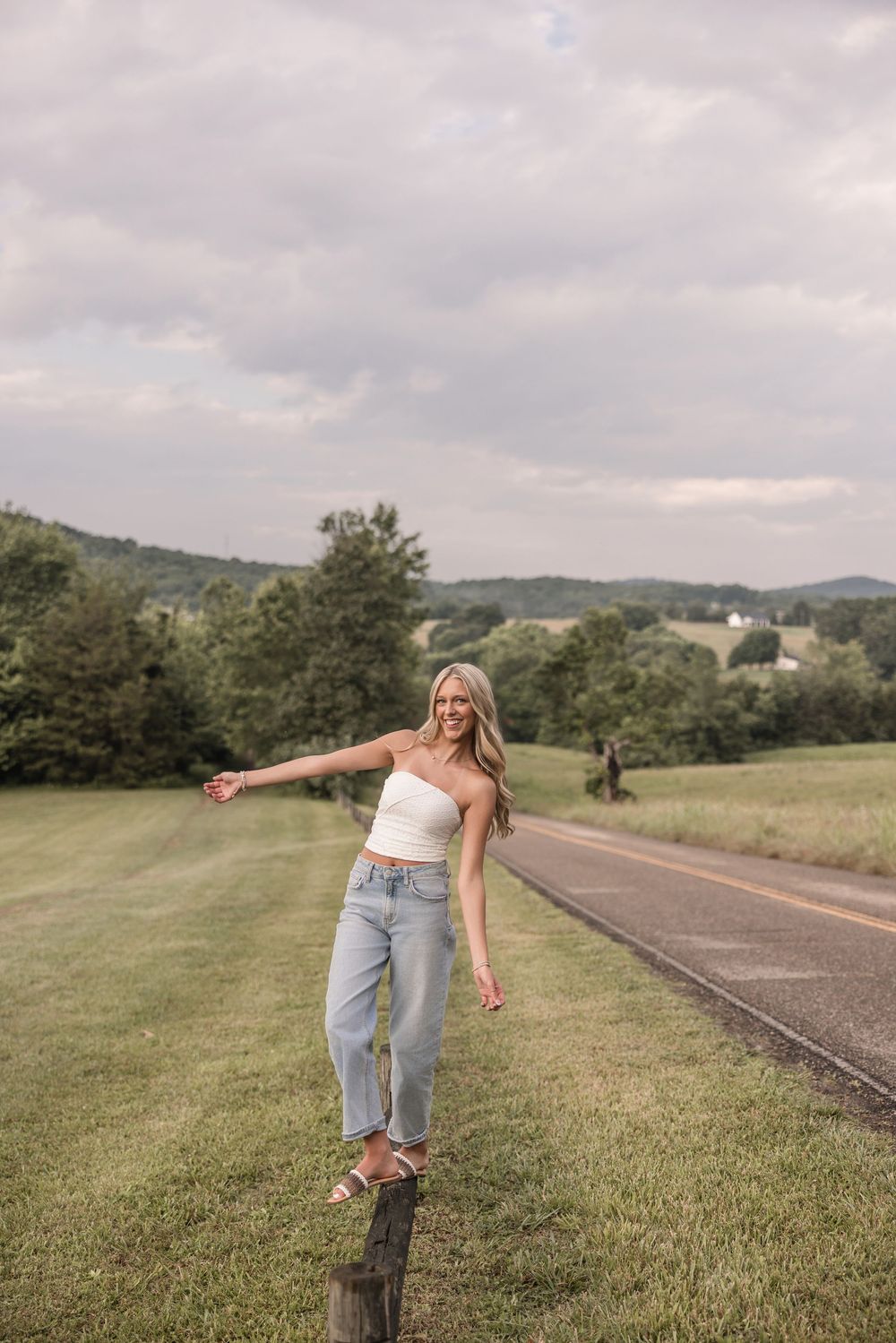 A person balances on a wooden fence post along a rural road surrounded by rolling green hills and cloudy skies.