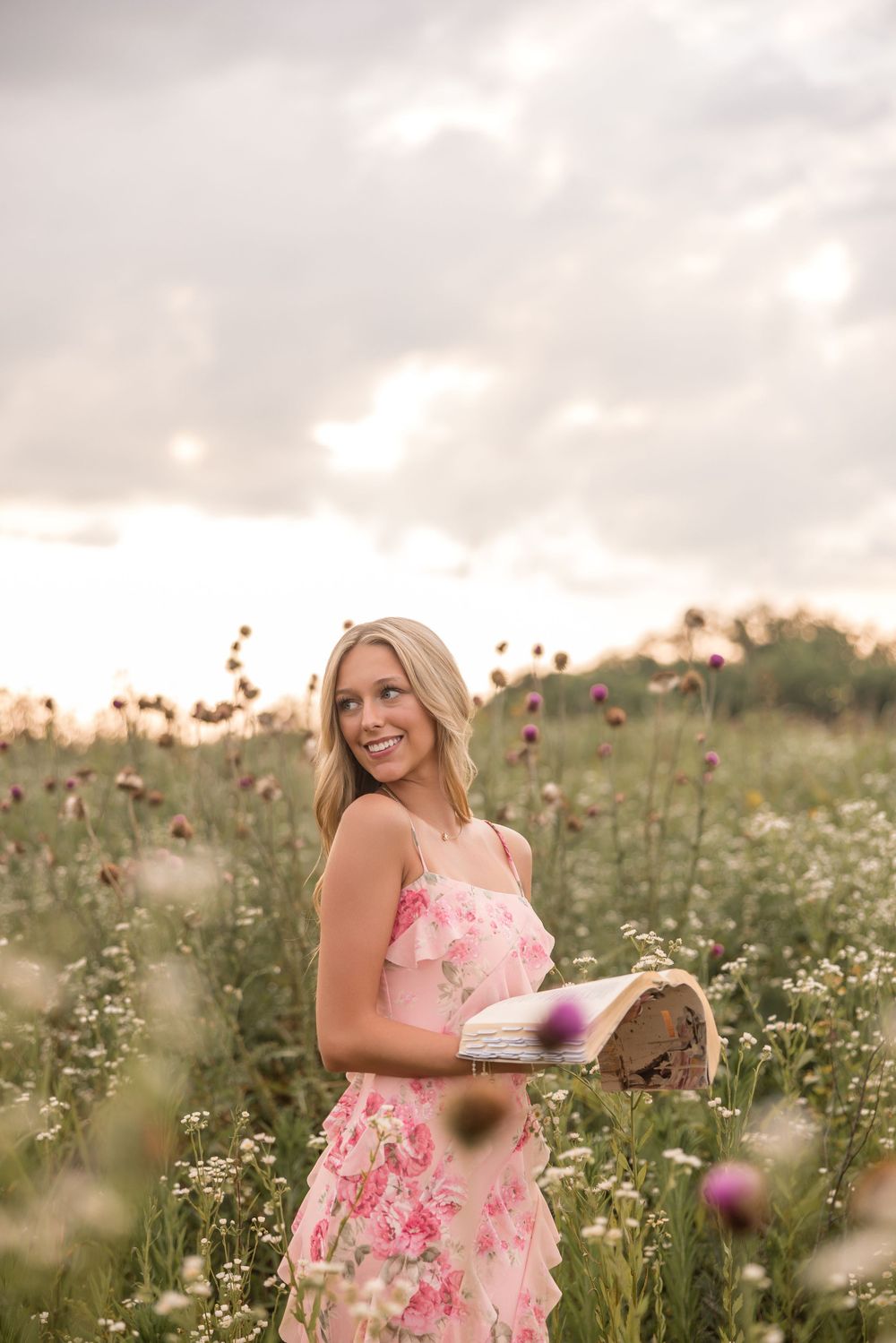 A dreamy outdoor portrait with a pink floral dress and book among wildflowers at golden hour.
