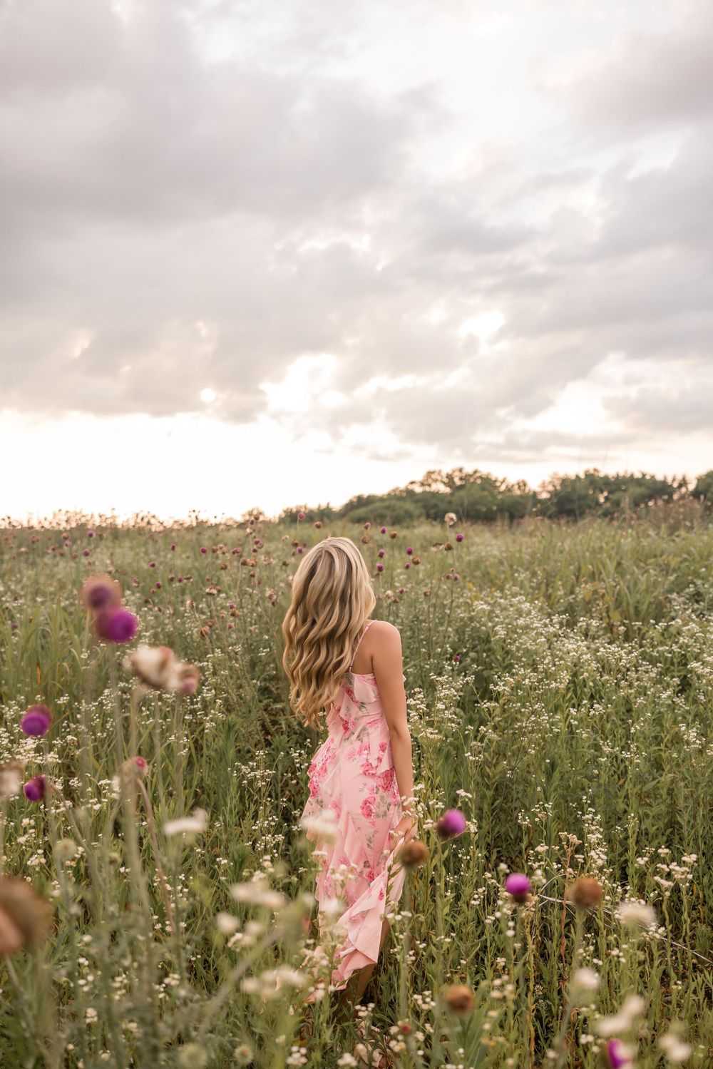 A figure in a pink floral dress stands among wildflowers in a meadow at sunset.