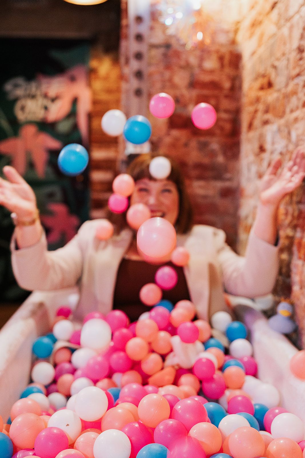 People celebrating at a party having fun in a ball pit filled with colourful pink, white and blue balls against brick walls.