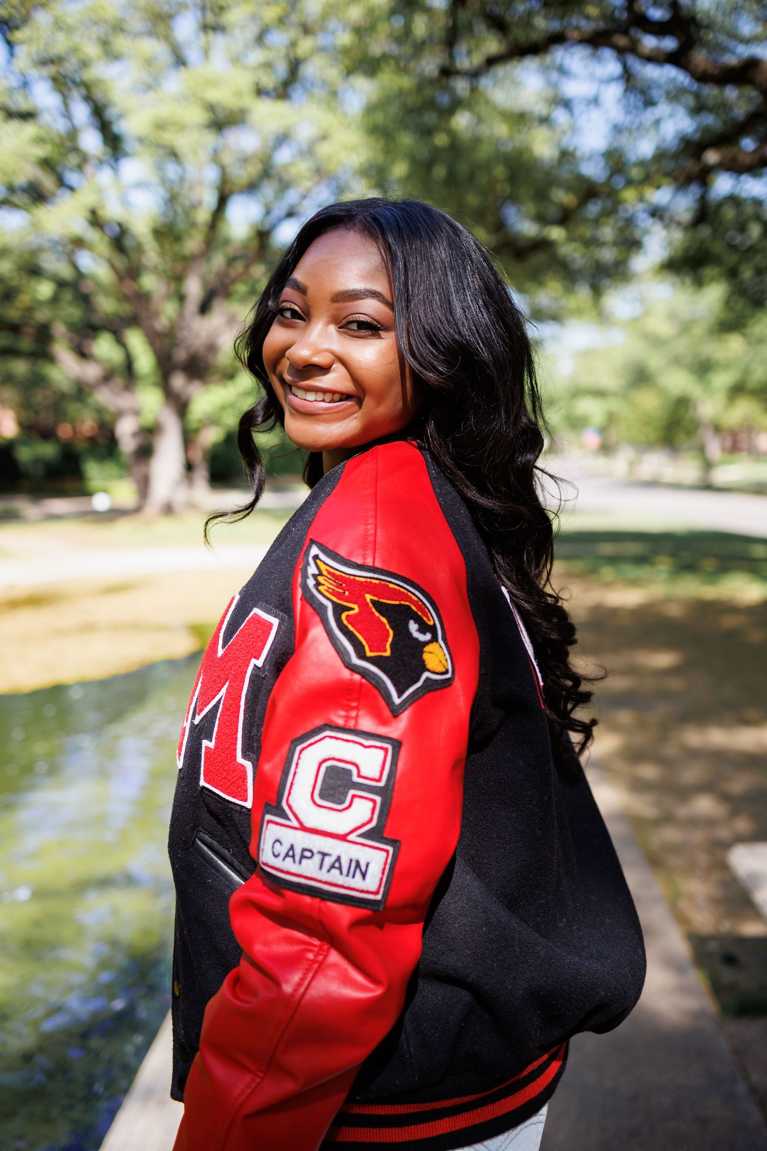 A student wearing a red and black Cardinals varsity letterman jacket with captain patch smiles outdoors on a sunny day graduation photo