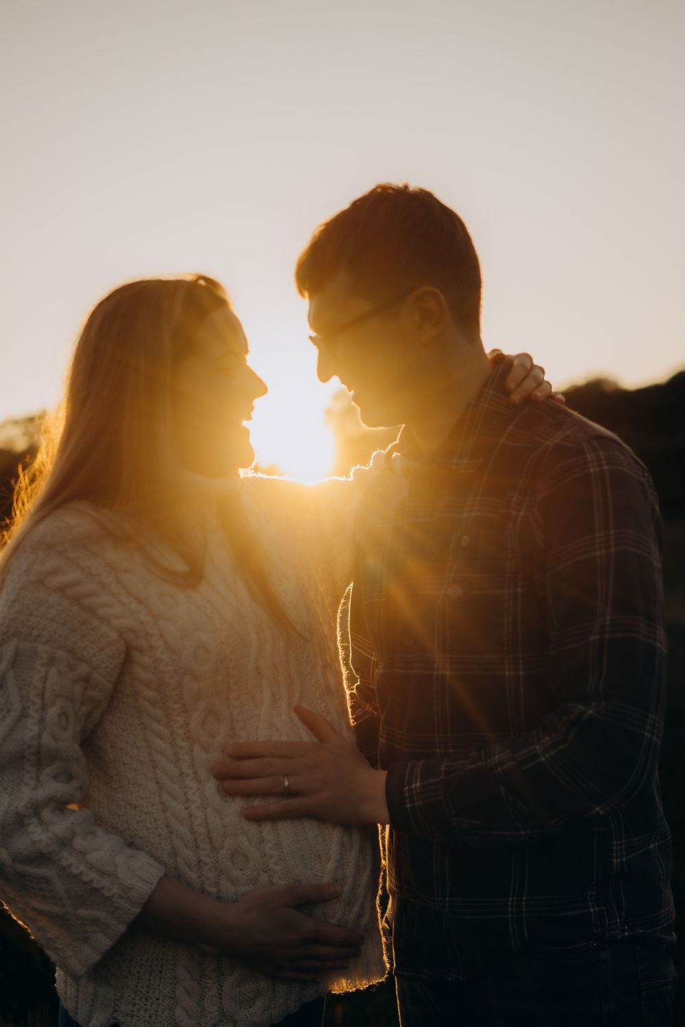 couple standing in sunset beams with man touching his wife's pregnant belly during maternity photoshoot in Stockholm