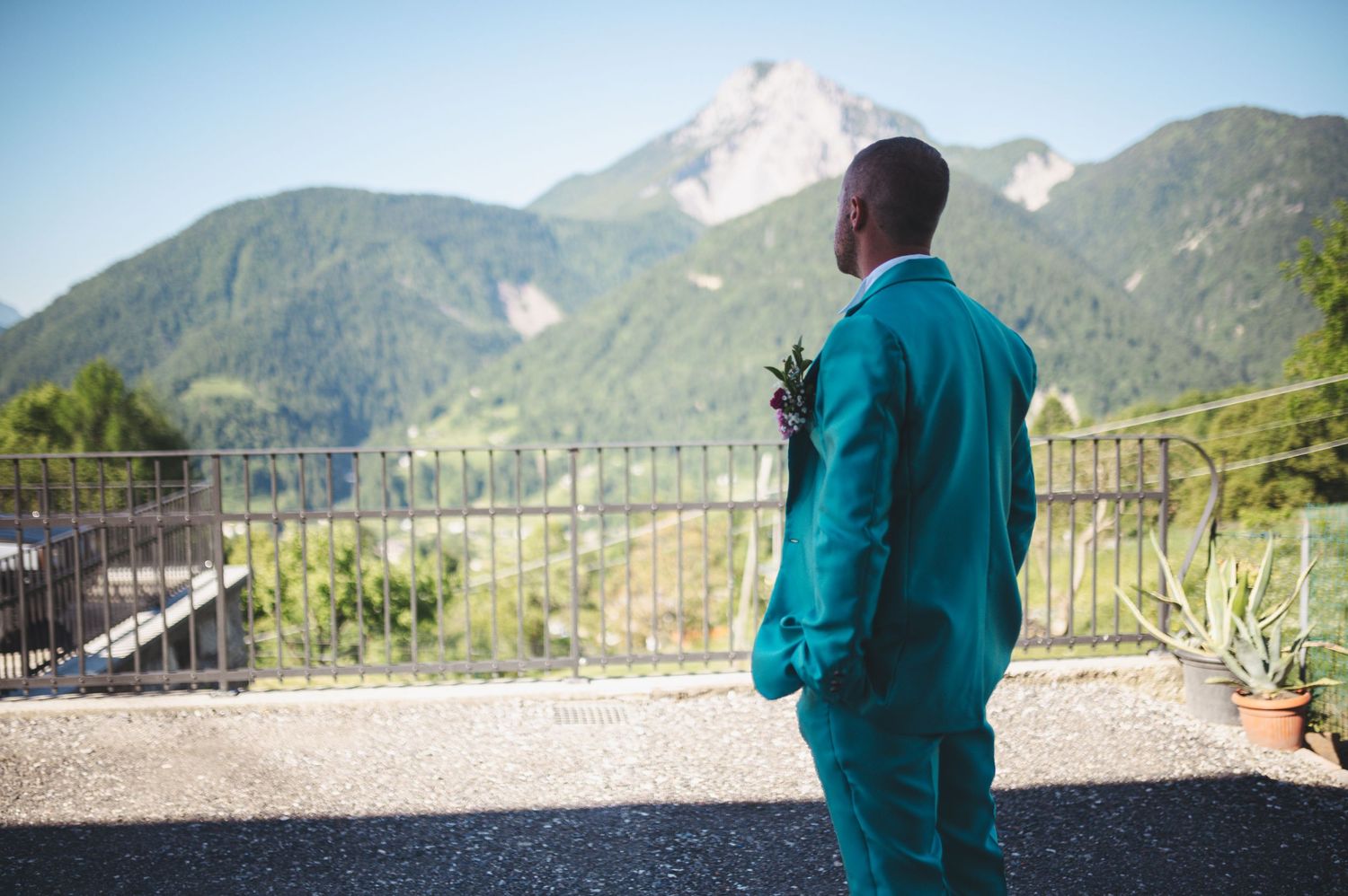 Person in teal suit stands silhouetted against mountain backdrop on outdoor terrace.