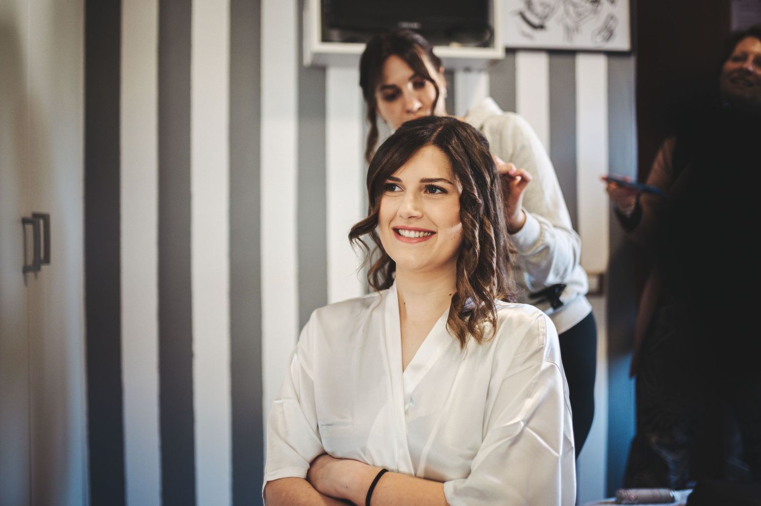 Woman in white robe smiles while getting hair styled in striped wall salon.