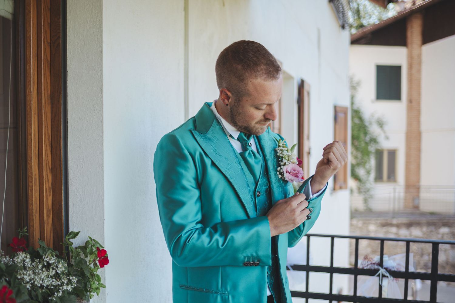 Person in bright turquoise suit adjusts boutonniere while standing on porch.