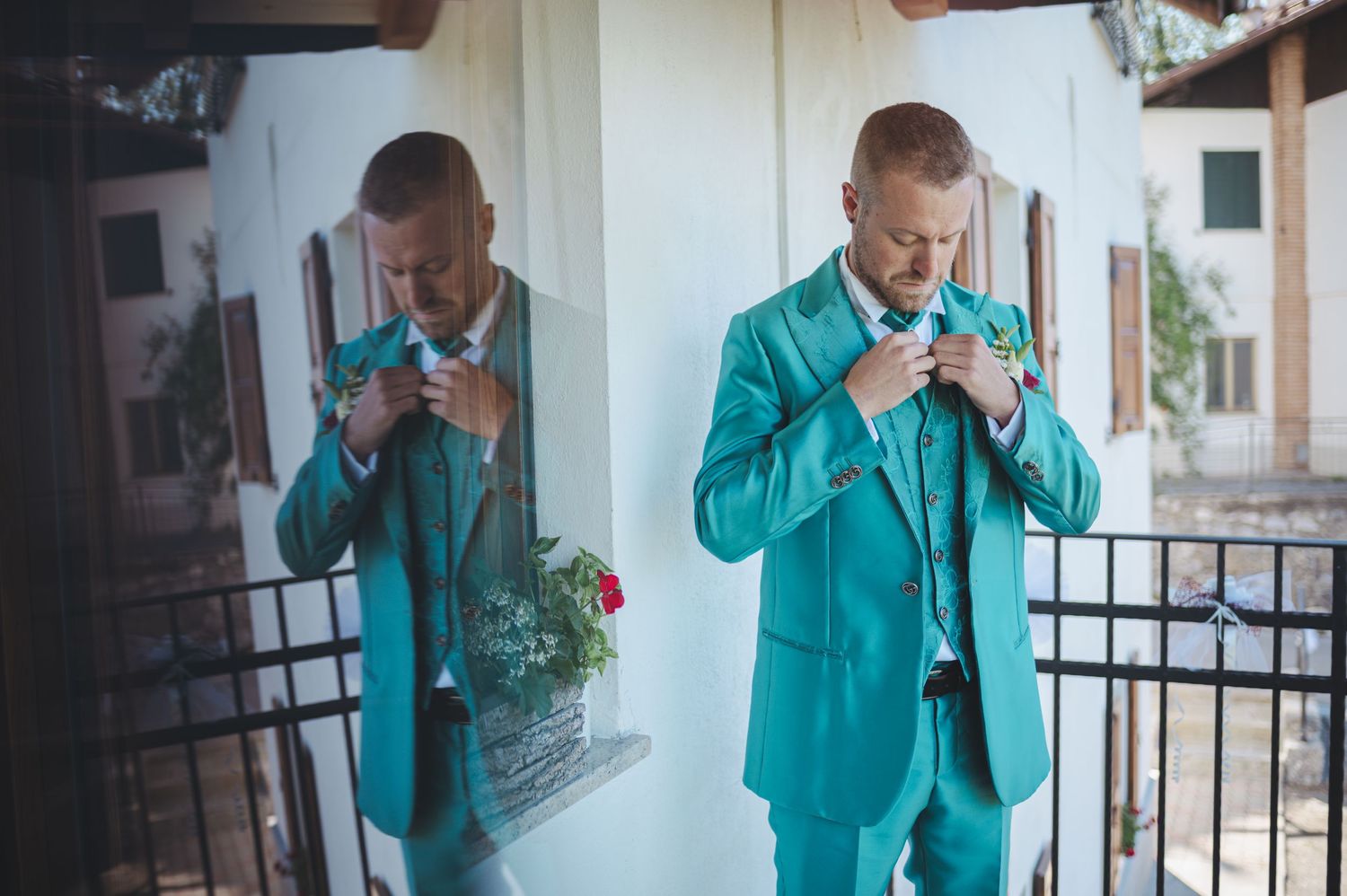 Groom in turquoise suit adjusts his bowtie while looking in mirror before wedding ceremony.