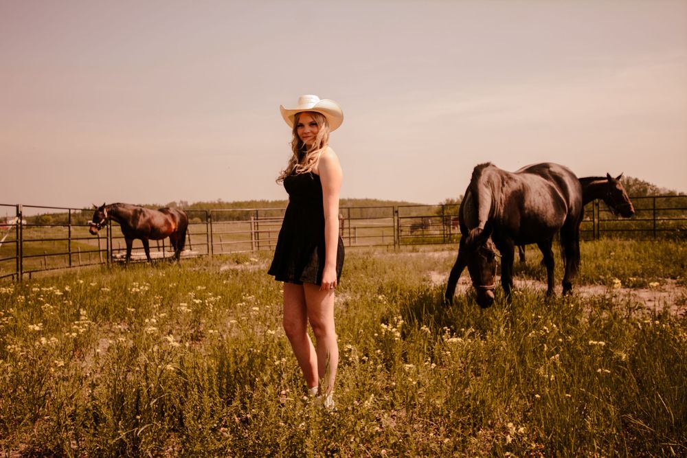 Woman in black dress and cowboy hat stands in sunlit field with grazing horses behind rustic wooden fence.