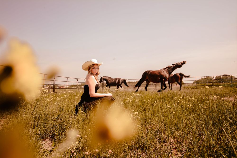 A photographer in a sundress captures horses running in a golden grassy field during sunset.