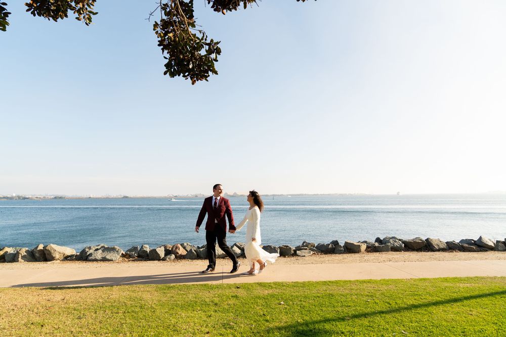 Couple takes a romantic stroll along a scenic oceanfront path with rocks and grass on a sunny day.