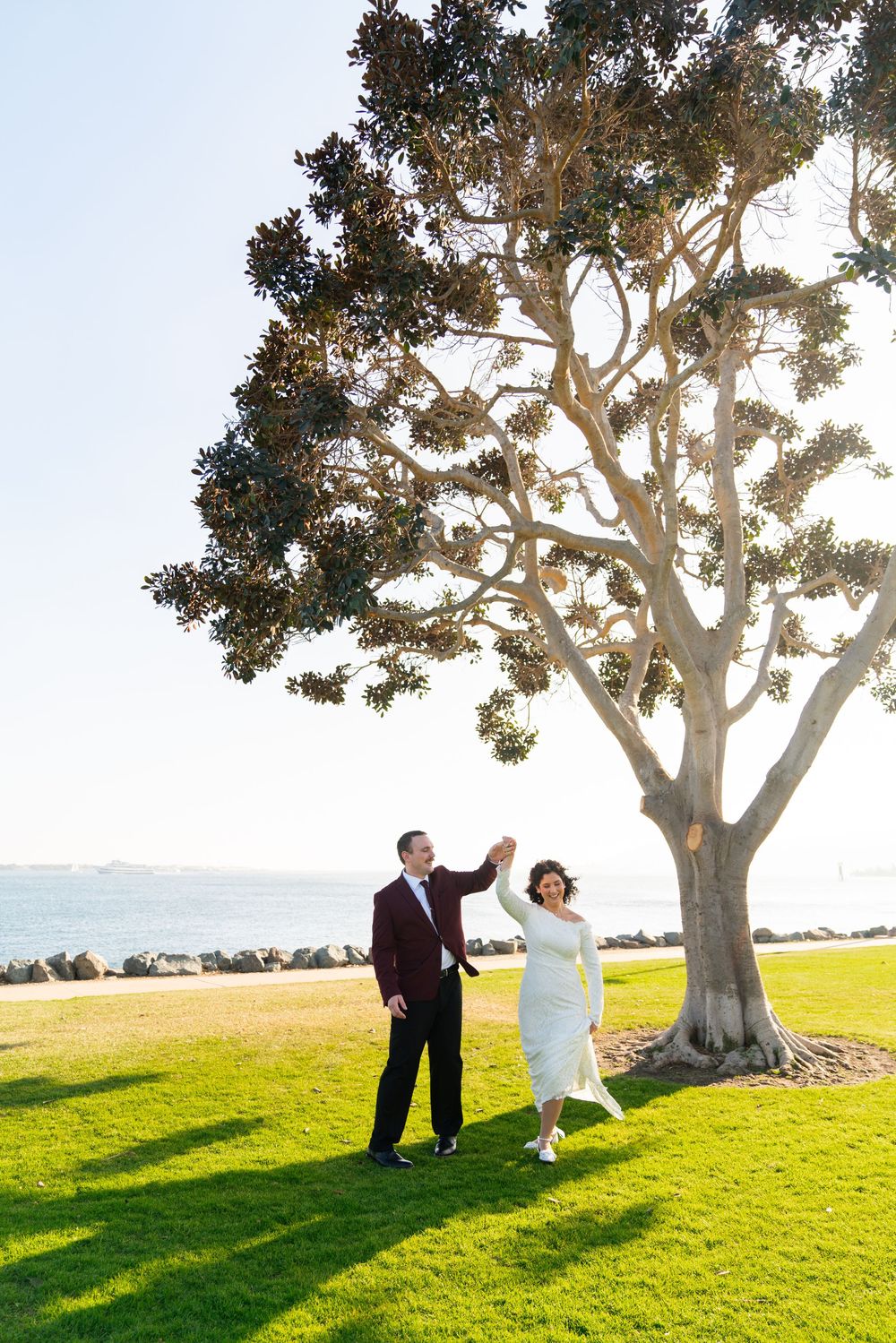 A couple dances joyfully on green grass near a large tree at sunset overlooking the ocean.