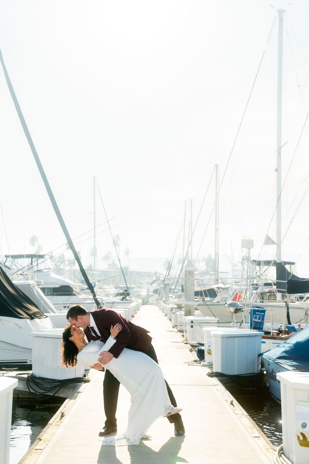A happy couple shares a romantic dip pose on a sunny marina dock surrounded by boats and sail masts.