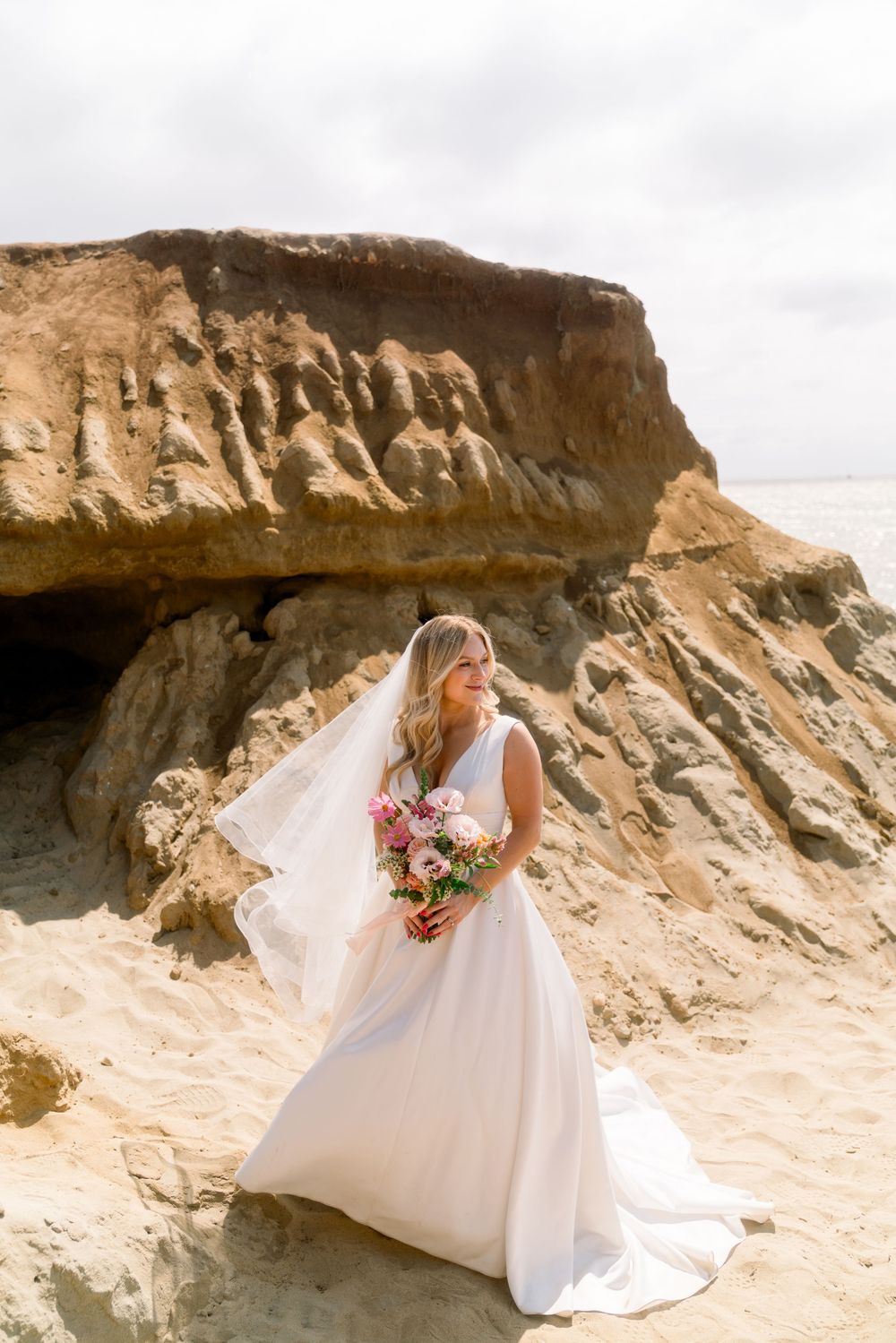 A bride in a flowing white dress holds a colorful bouquet while standing on dramatic coastal cliffs under cloudy skies.