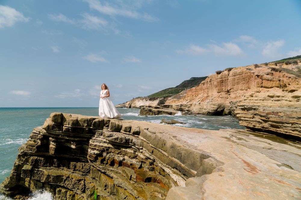 A white-clad figure stands on rugged coastal cliffs overlooking the turquoise ocean against a blue sky.