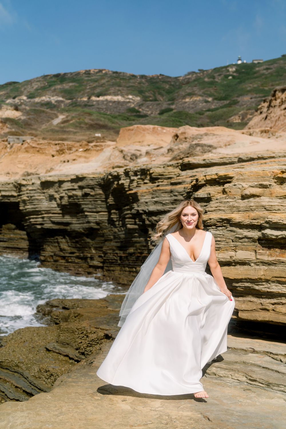 A wedding dress flows elegantly against dramatic coastal cliffs and crashing waves at a scenic oceanside location.