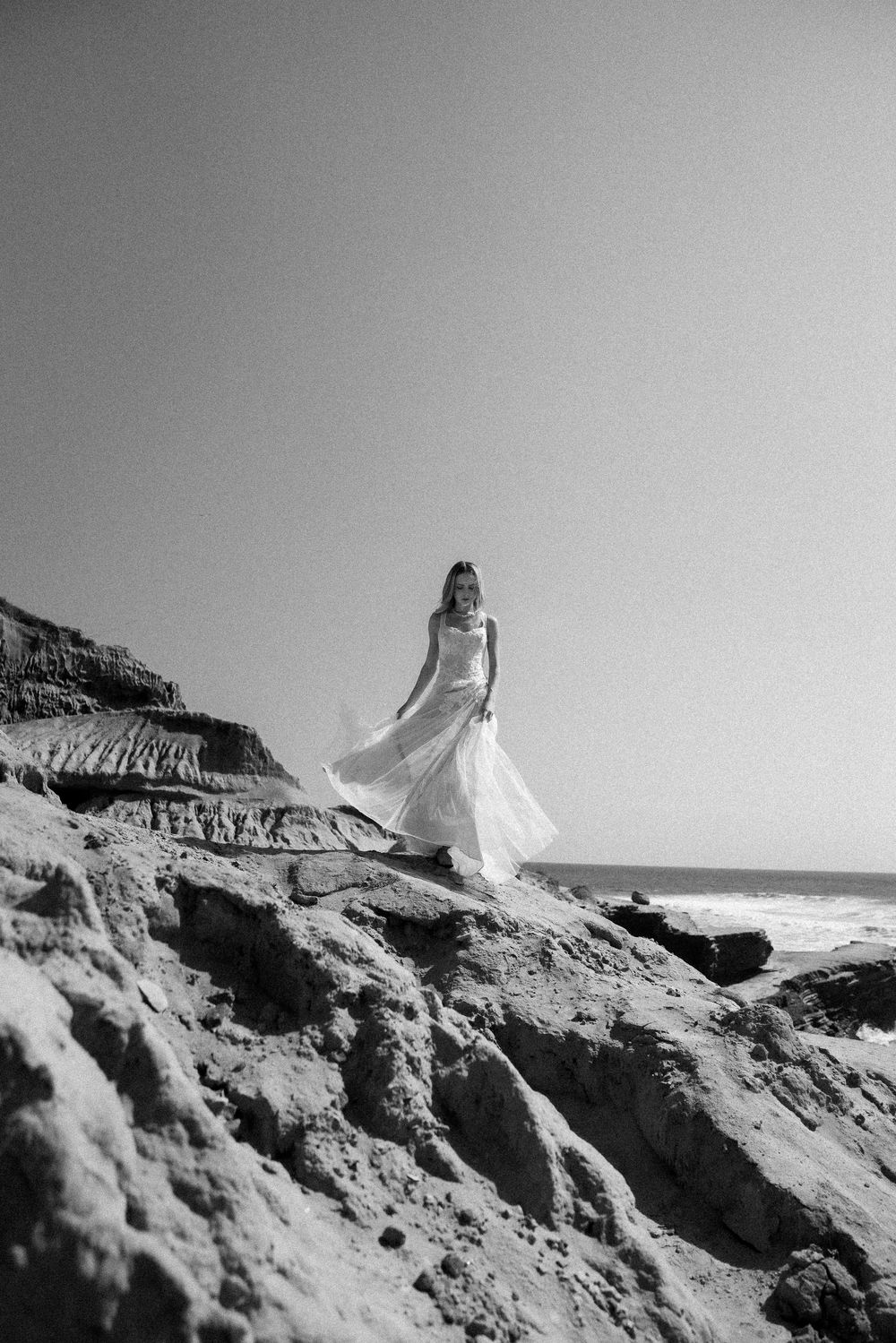 Black and white photo of an elegant figure in a flowing white dress standing on dramatic coastal cliffs by the ocean.