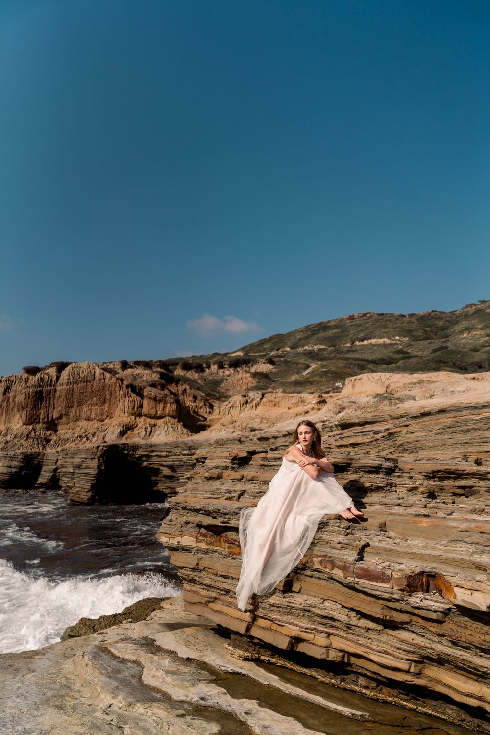Woman in flowing pink dress poses elegantly on rocky coastal cliffs as waves crash against the shore at sunset.