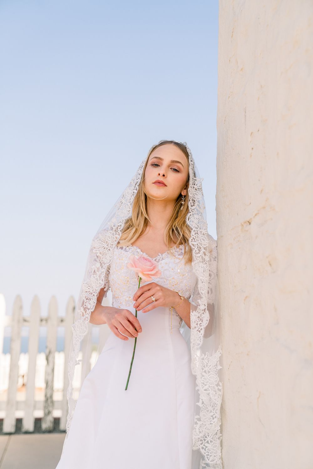 A figure in a white lace wedding dress and veil stands against a wall holding a single pink rose on a sunny day.