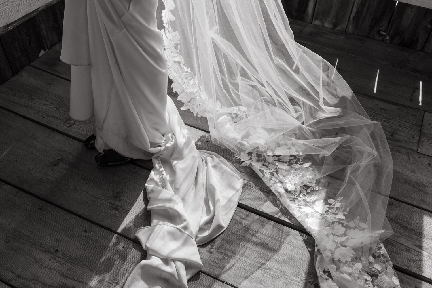 Black and white photograph showing intricate lace details of a wedding dress train flowing across a wooden floor.
