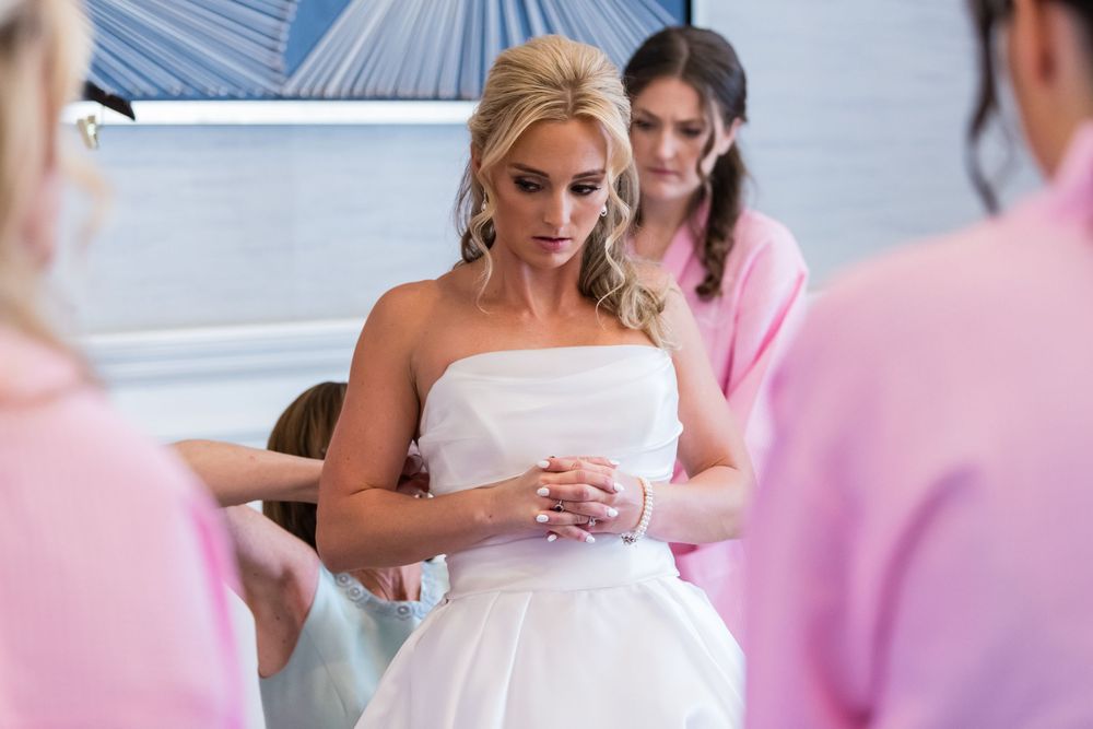 Person in a strapless white wedding gown looks at phone while others in pink surround them during preparation.