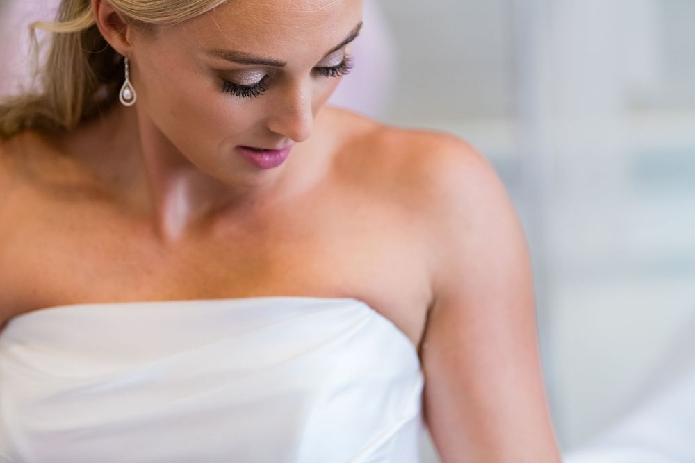 Young bride in a strapless white wedding gown looking down with elegant makeup and blonde hair.