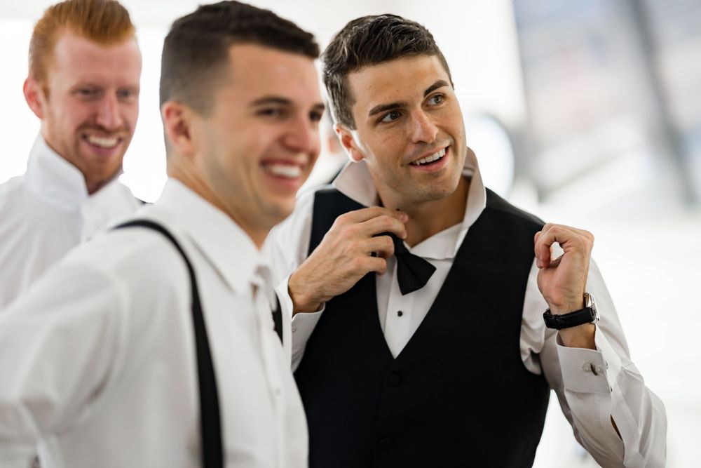 Groomsmen in tuxedos share a candid moment and laugh together while adjusting bowties before wedding ceremony.