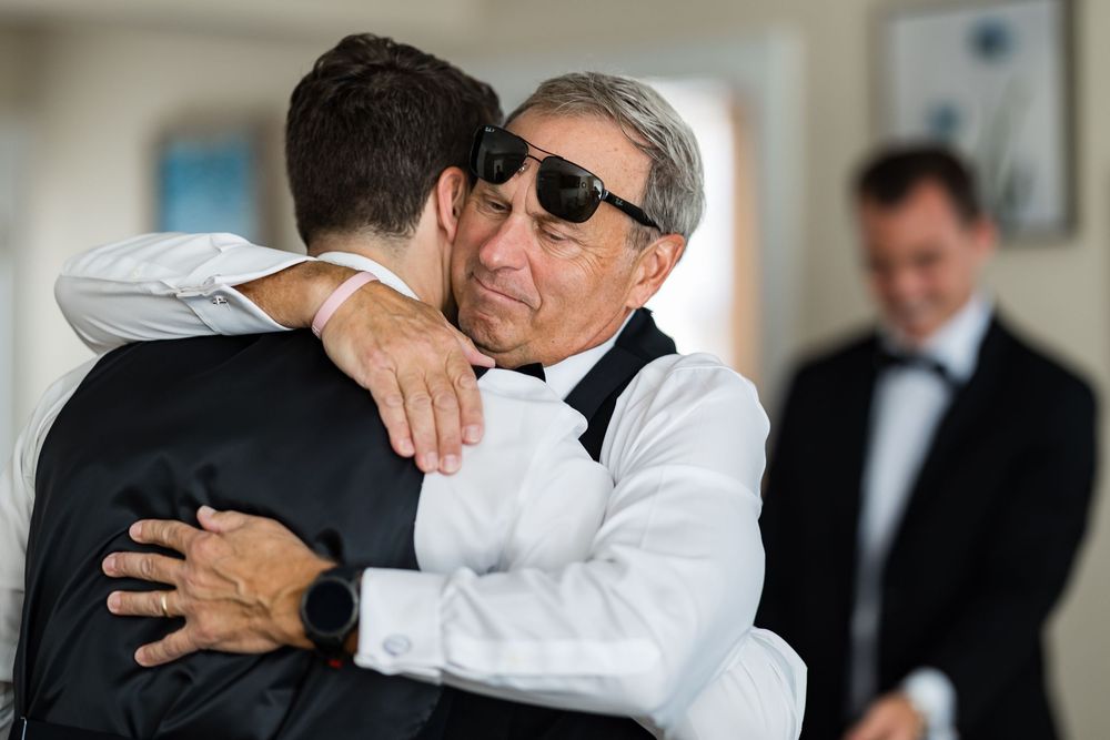 Two people dressed in formal attire share an emotional embrace while another person looks on in the background.