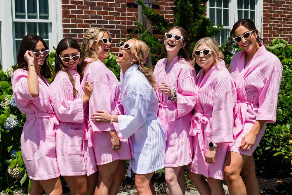 Group of people wearing matching pink and lavender robes laugh and smile together at outdoor bridal party celebration.
