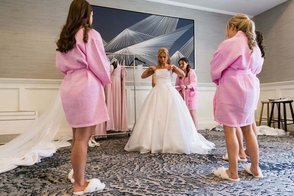 Bridesmaids in pink robes help adjust the wedding dress and veil of a bride in a luxurious hotel room.