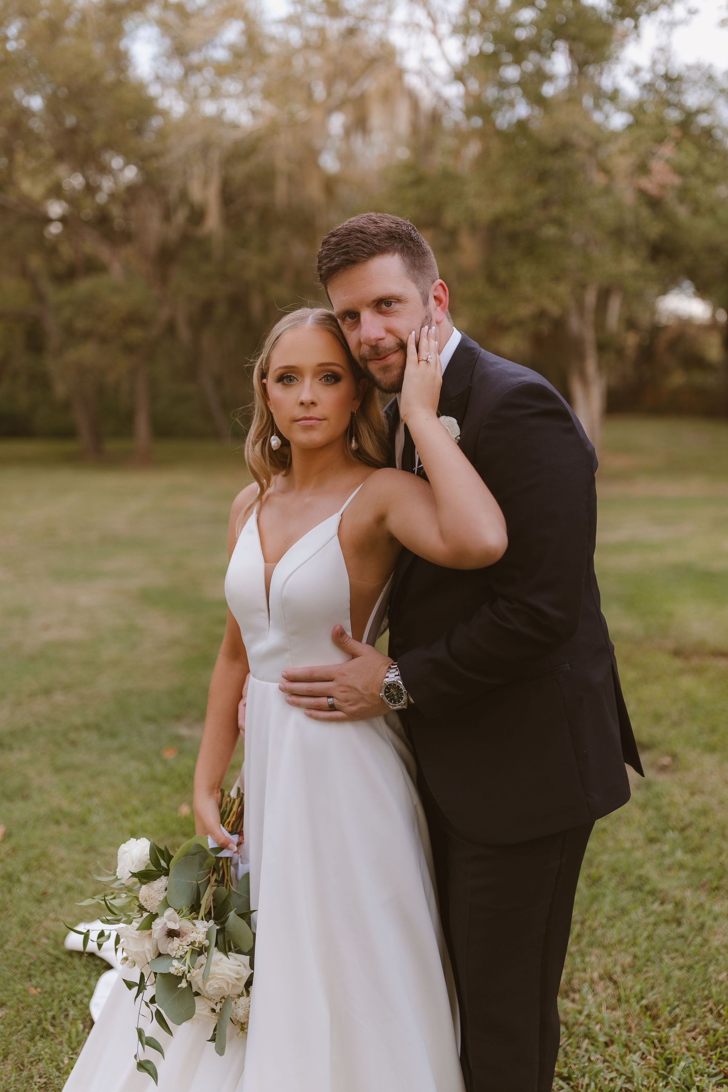 Bride in elegant white dress and groom in black tuxedo embrace outdoors during sunset wedding photo session.
