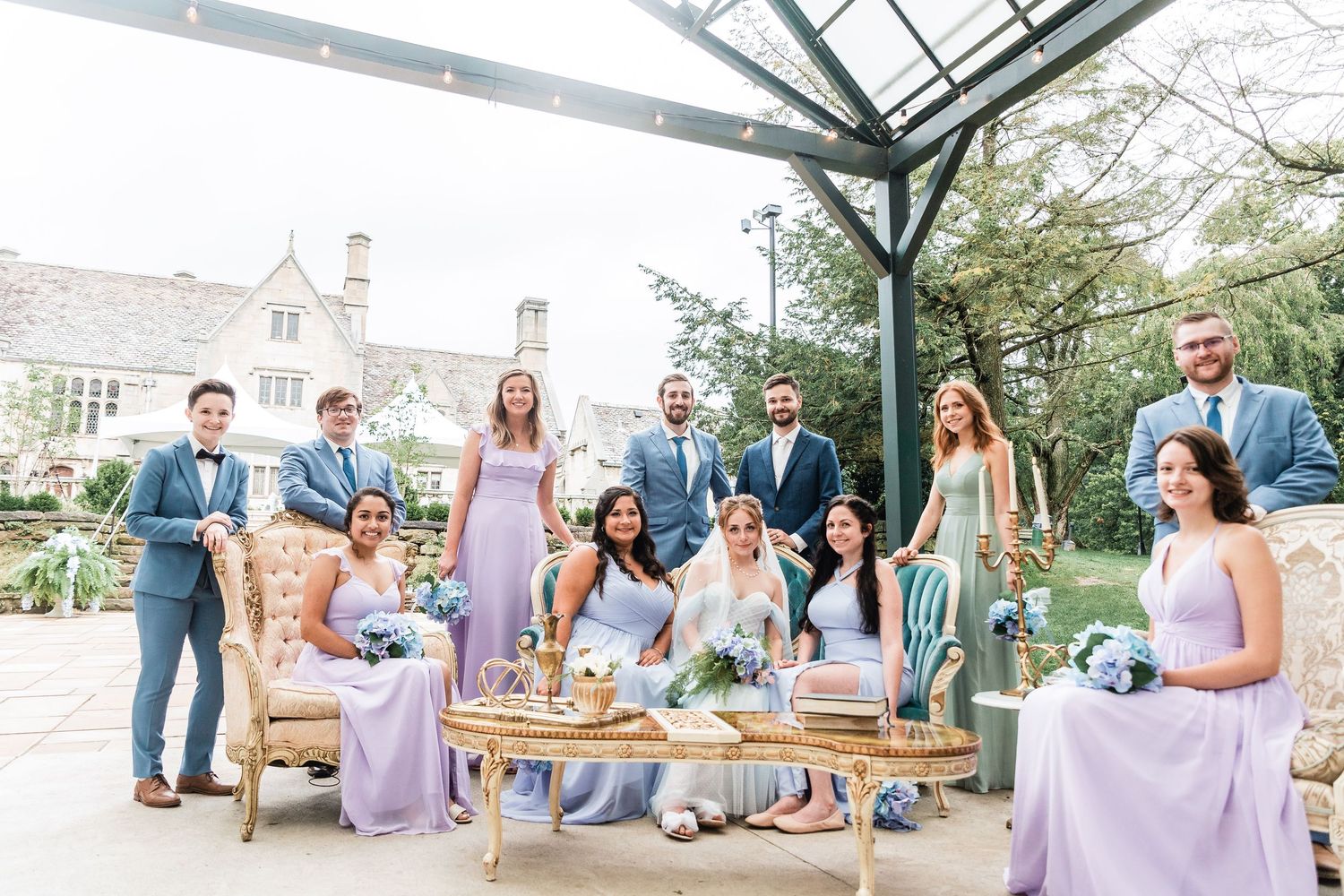 Wedding party in light purple bridesmaid dresses and blue suits gather on outdoor patio with rustic furniture.
