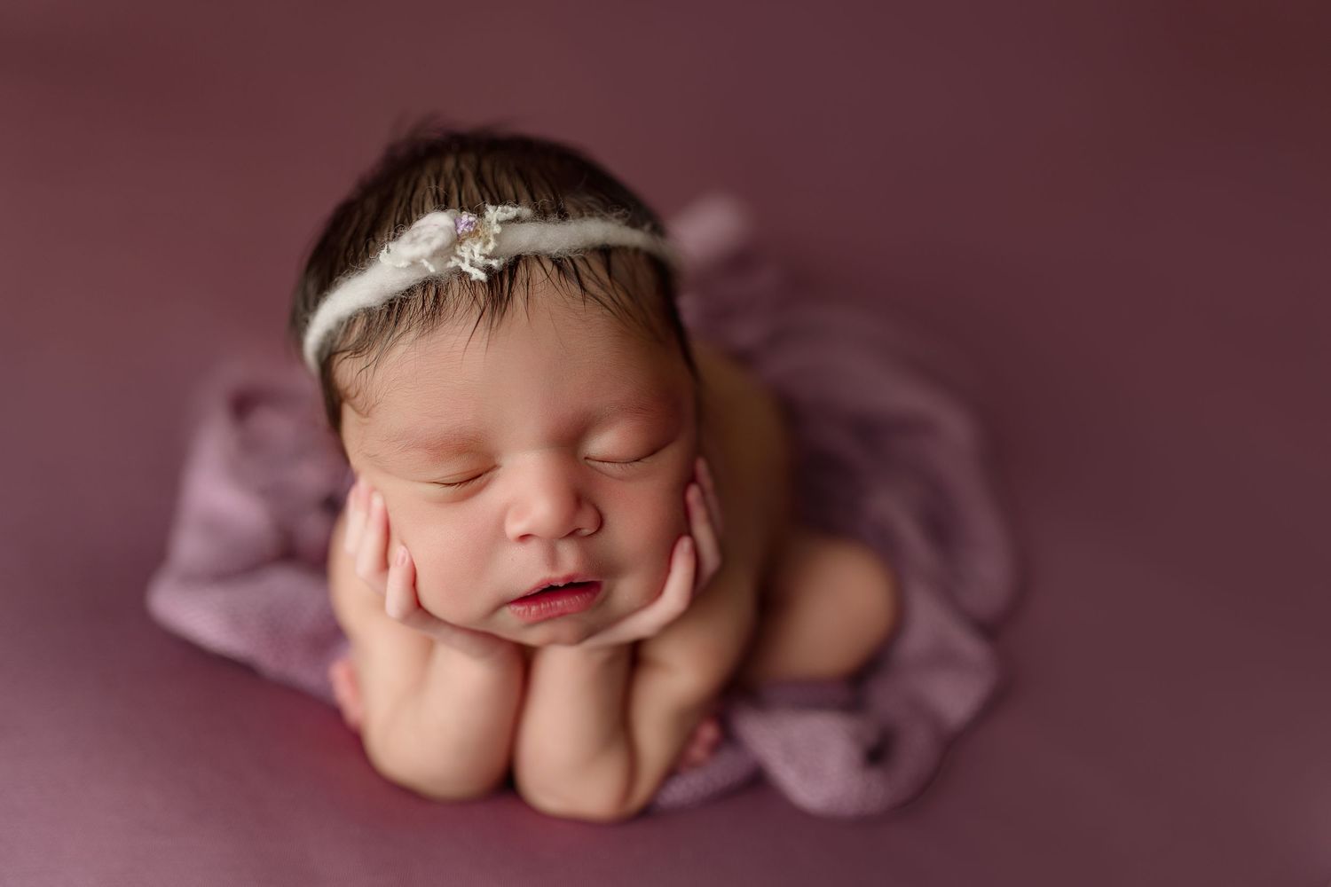 Sleeping newborn on pink fabric in frog pose wearing delicate white headband during professional photo session.