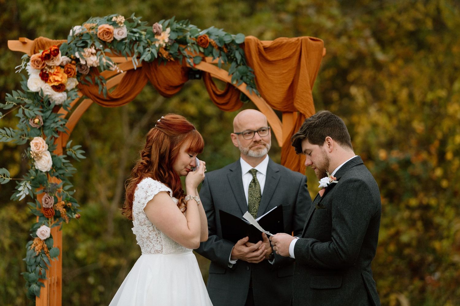 A couple exchanges rings during an outdoor autumn wedding ceremony beneath a floral-adorned wooden arch.