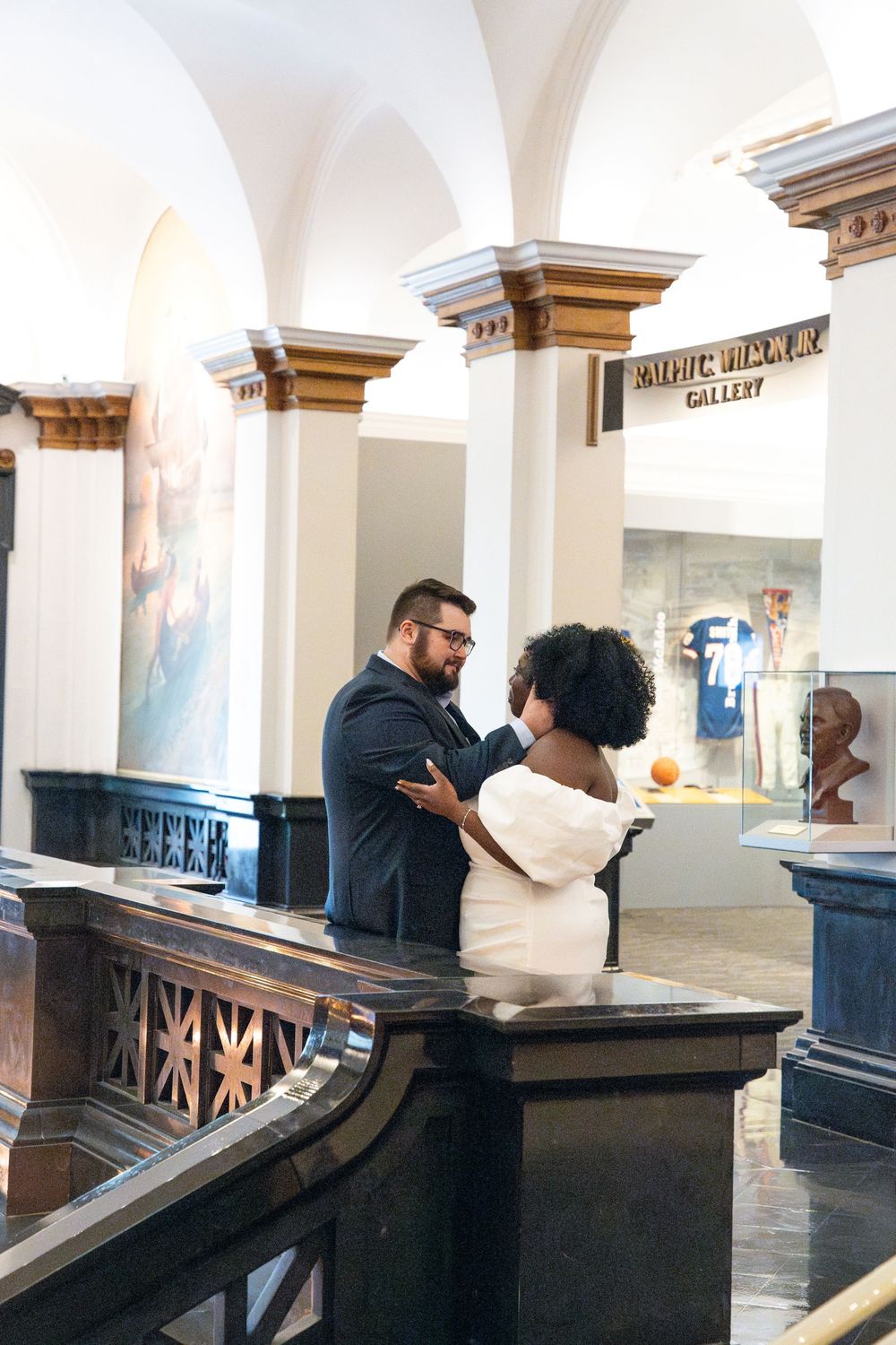 Two people having an animated conversation while standing under white arched columns in an elegant indoor gallery space.