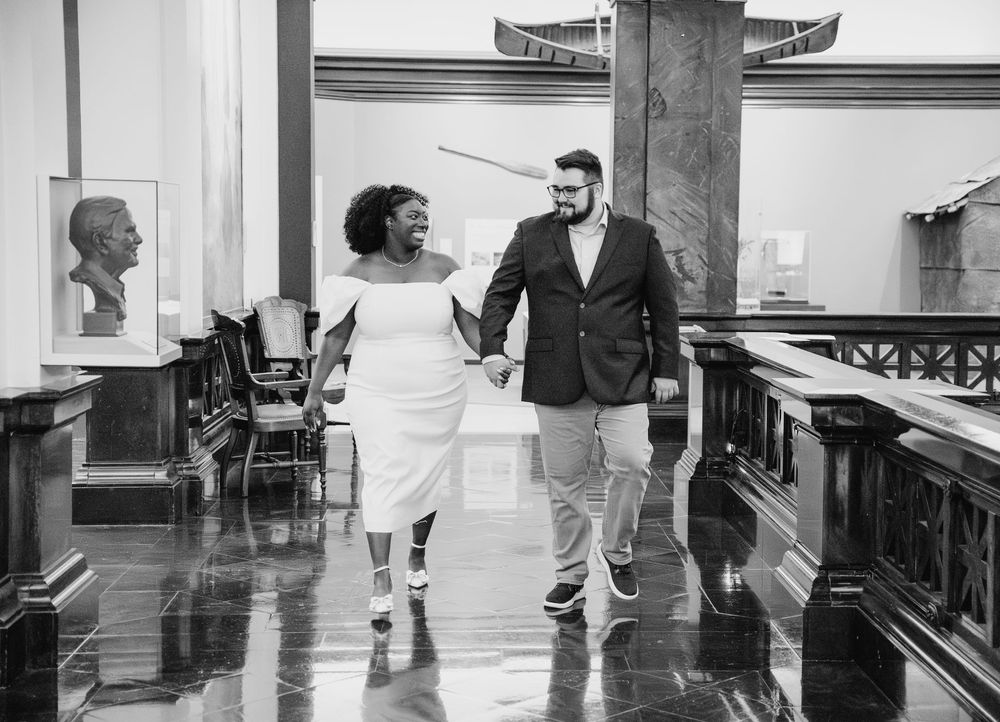 A joyful couple walks together in a dramatic black and white photo inside an ornate vintage building interior.