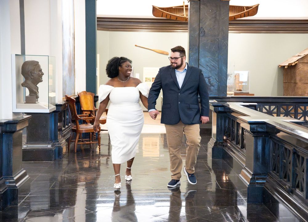 A couple walks hand-in-hand through an elegant hallway with art displays and wooden details.