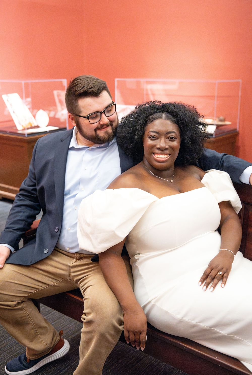 A couple in formal attire smile together while sitting on a burgundy couch, the bride wearing an elegant white dress.