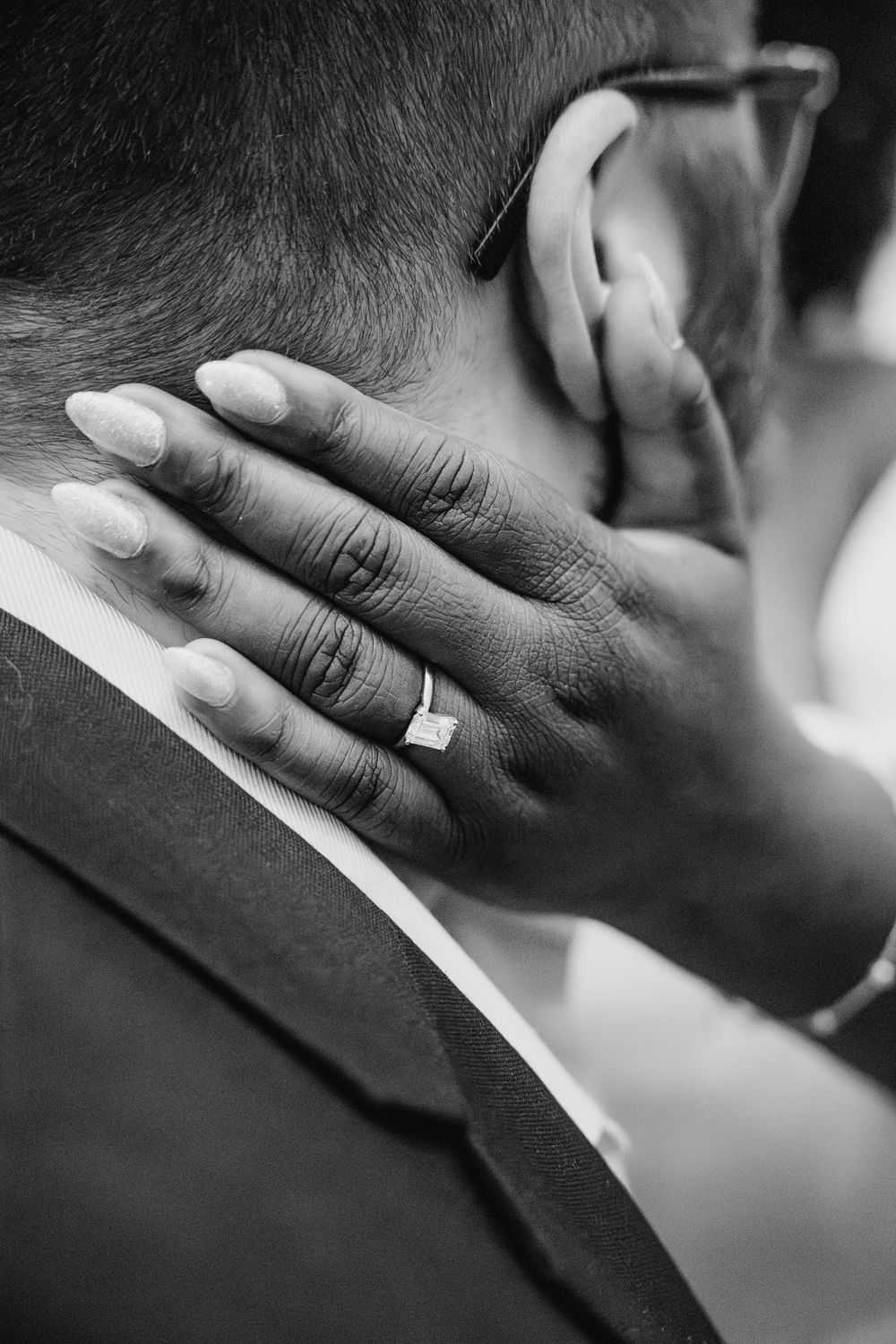 A black and white close-up photo of a wedding ring on a hand resting against a formal suit.