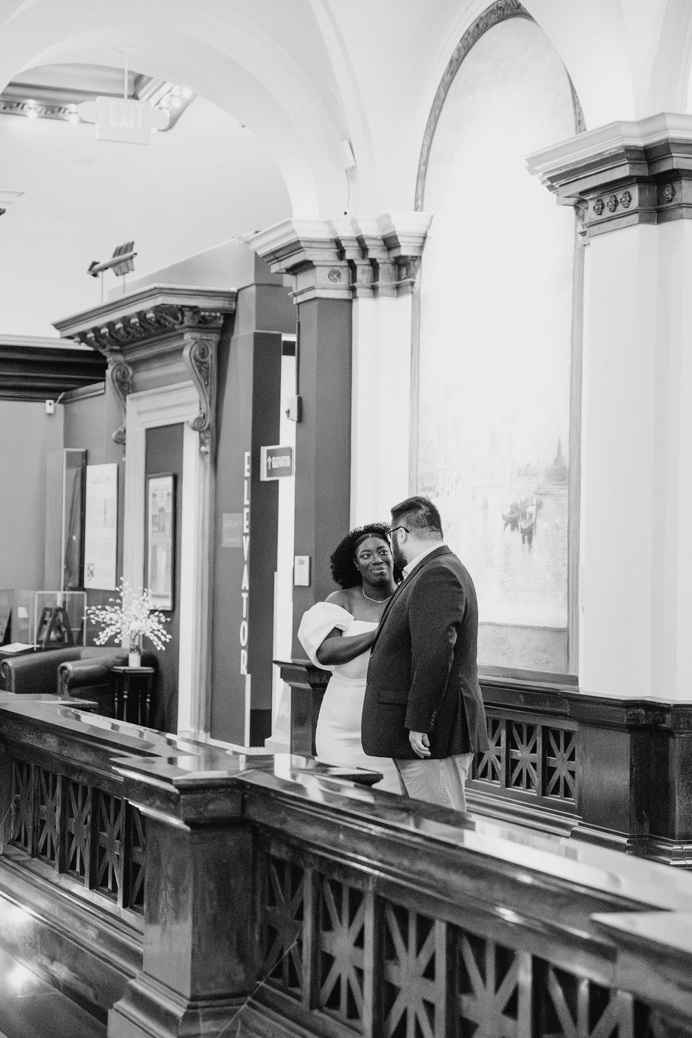 Black and white photo of a couple sharing a romantic moment on an elegant balcony with ornate architectural details.