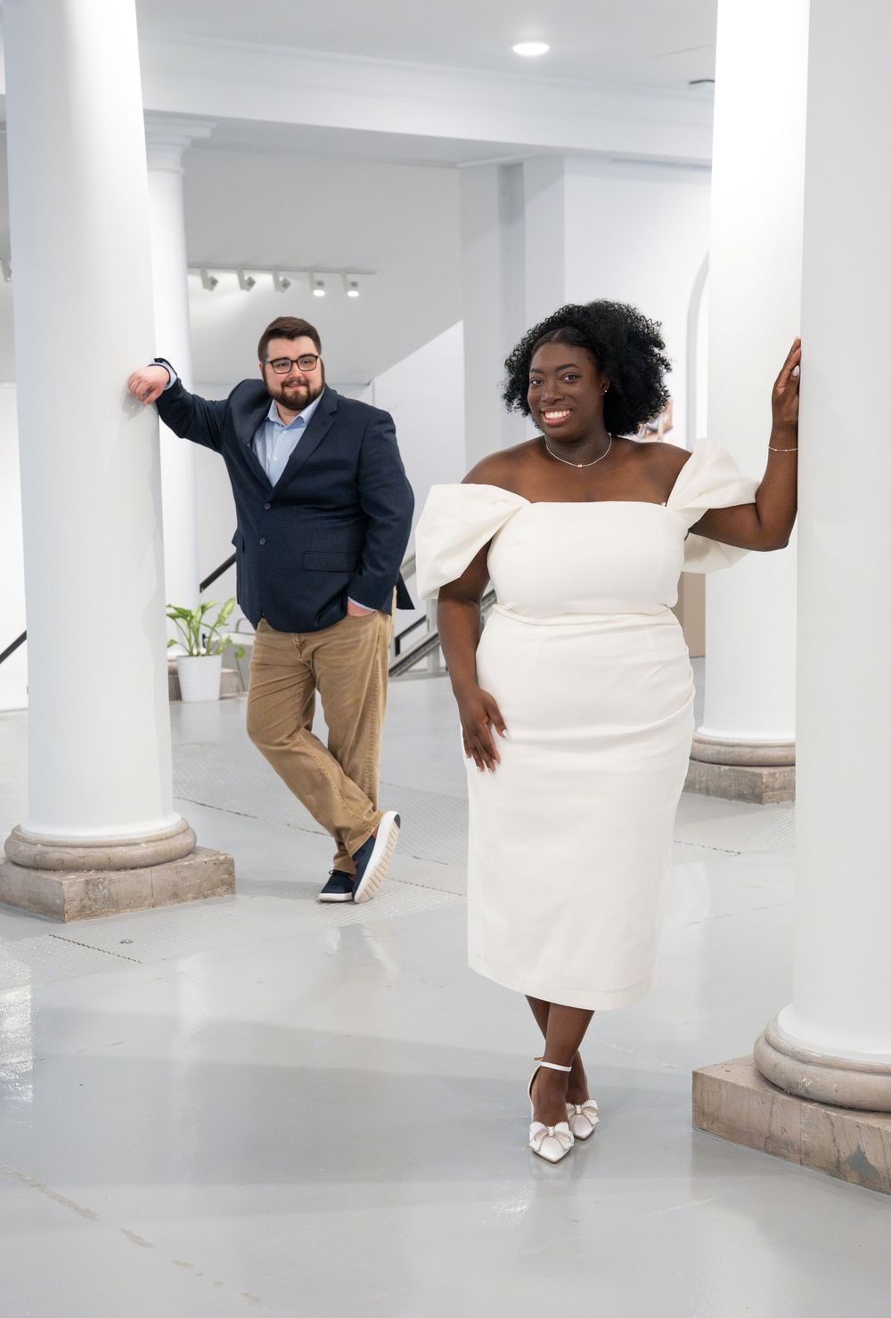 A couple poses playfully between white columns in an elegant indoor setting with modern decor.