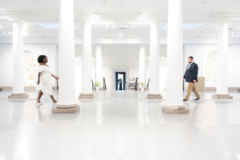 Two people walk through a bright white modern interior space with columns and wooden stepping platforms.