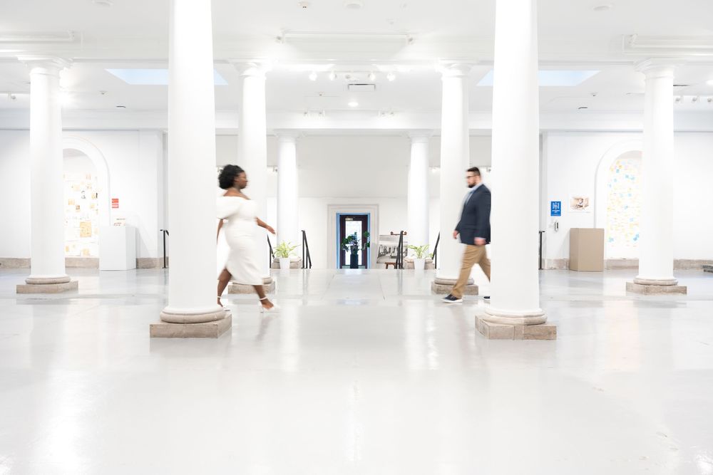 Two people stand apart in a bright minimalist white hallway with columned architecture.