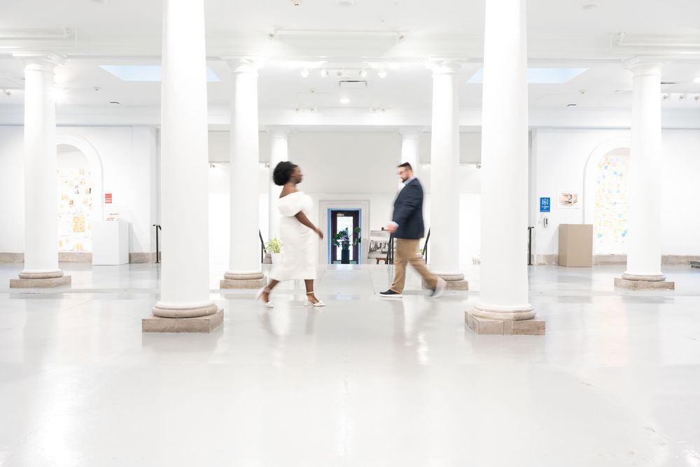 Two people walking through a bright white modern museum gallery space with pillars and artwork on display.