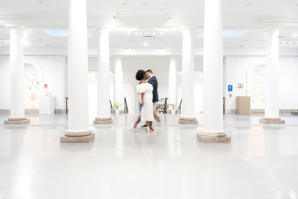 Two people embrace in an elegant white hallway with classical columns and high ceilings.